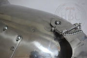Close-up of a polished metallic medieval-style helmet or armor plate with rivets and a chain fastener, shown against a light neutral background.
