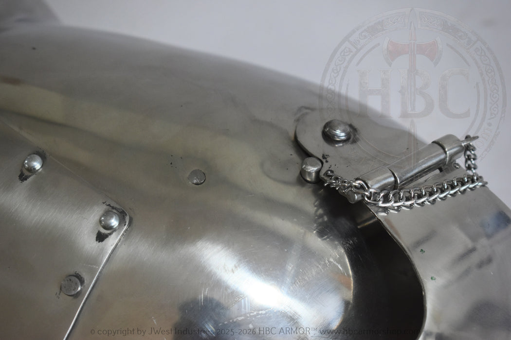 Close-up of a polished metallic medieval-style helmet or armor plate with rivets and a chain fastener, shown against a light neutral background.