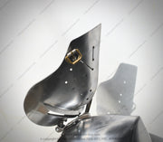 A close-up of a metallic armor plate or shield component with a polished steel finish, featuring pre-drilled holes and a small brass-colored hardware detail, set against a light gray background with a prominent cast shadow.