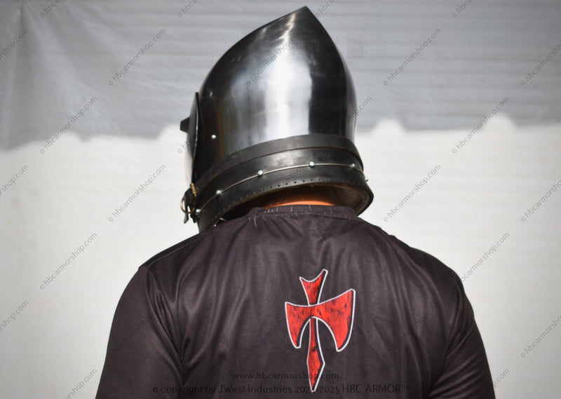 A person viewed from behind wearing a polished steel medieval-style helmet and a black tunic with a red Templar cross emblem on the back, set against a white draped backdrop.