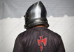 A person viewed from behind wearing a polished steel medieval-style helmet and a black tunic with a red Templar cross emblem on the back, set against a white draped backdrop.