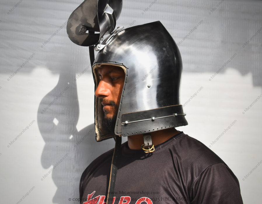 A man wearing a polished steel medieval-style helmet with a visor and curved top ornament, shown in profile against a light gray wall.