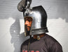 A man wearing a polished steel medieval-style helmet with a visor and curved top ornament, shown in profile against a light gray wall.