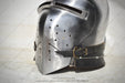 A medieval steel helmet with a visor and a dark leather chin strap, photographed on a light gray textured background.