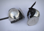 Two medieval-style steel helmet visors with black leather straps, displayed on a light gray background. One visor is shown closed and the other is shown open to display the internal structure and spike.