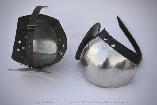 Two medieval-style metal helmets displayed side by side on a plain light background, one showing the interior with a leather strap and the other showing the exterior with a leather visor and rear strap.