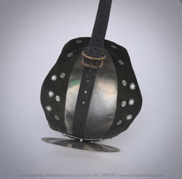 A medieval-style steel and leather visor or helmet component, featuring a polished steel plate, a black leather strap with a brass buckle, and a circular steel base, photographed against a light gray background.