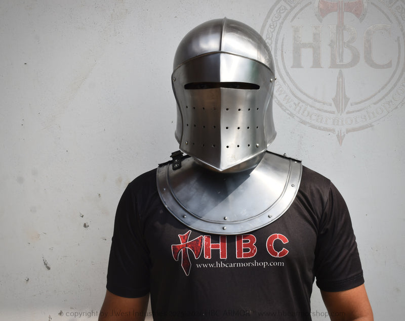 A person wearing a medieval steel helmet with a visor and matching breastplate armor, standing against a light gray wall. The person is wearing a black t-shirt with a red and white HBC logo.