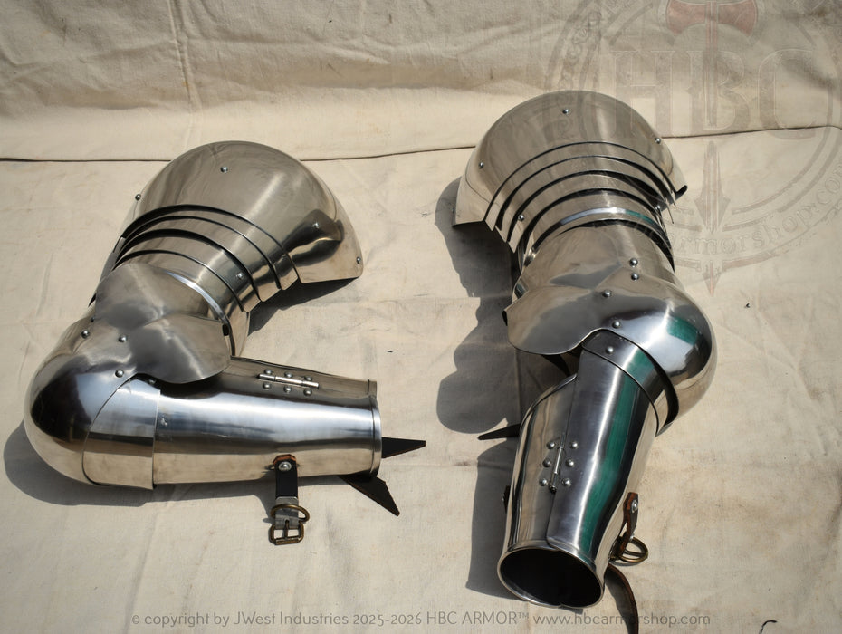A pair of polished steel medieval chainmail gauntlets with curved, articulated sleeves, displayed on a light beige fabric backdrop.