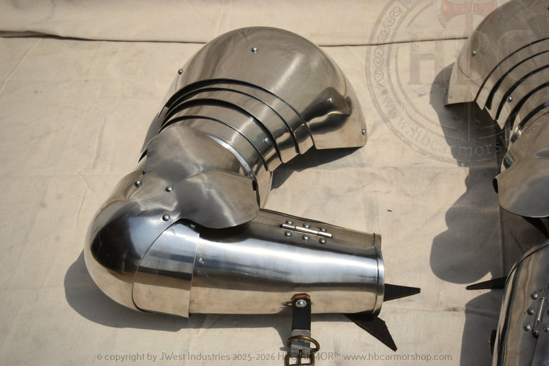 A polished steel medieval-style armor breastplate or guard with rivets and a pointed tip, photographed on a light fabric surface alongside other matching metal armor pieces.