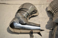 A polished steel medieval-style armor breastplate or guard with rivets and a pointed tip, photographed on a light fabric surface alongside other matching metal armor pieces.