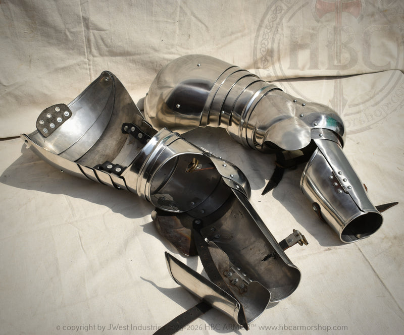 A pair of medieval steel plate armor leg guards and shin guards displayed on a light beige fabric backdrop.