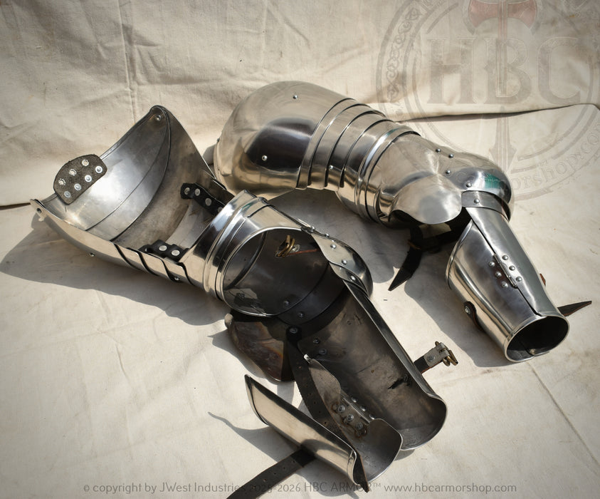 A pair of medieval steel plate armor leg guards and shin guards displayed on a light beige fabric backdrop.