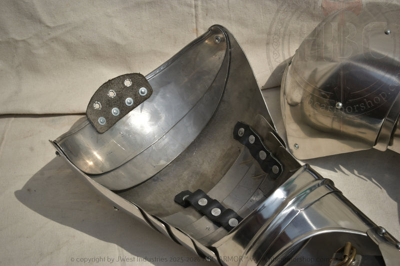 Close-up of a polished steel medieval breastplate or armor plate with rivets and straps, shown on a beige fabric background with part of a matching helmet visible.