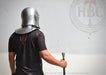 A person wearing a silver metal helmet and shoulder armor stands with their back to the camera, holding a sword hilt against a light gray wall. The person is wearing a black t-shirt with a red graphic on the back.