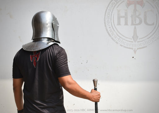 A person wearing a silver metal helmet and shoulder armor stands with their back to the camera, holding a sword hilt against a light gray wall. The person is wearing a black t-shirt with a red graphic on the back.
