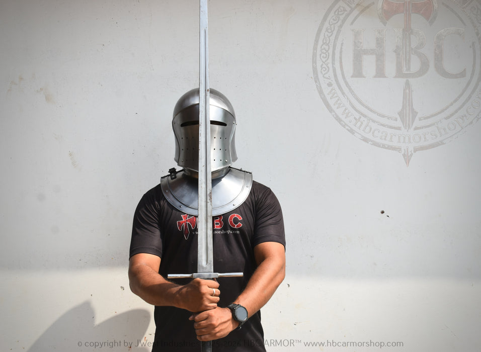 A person wearing a medieval-style steel helmet and chainmail armor stands holding a long sword upright in front of a light gray wall. The person is wearing a black t-shirt with a red and white logo.