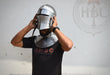 A man wearing a polished steel medieval knight helmet and matching steel neck guard, adjusting the helmet with both hands. He is wearing a black t-shirt with red and white 'HBC' branding.