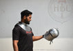 A man wearing a black t-shirt and a silver steel neck guard holds a matching silver steel medieval helmet in front of a light gray wall with a faint HBC Armor logo.
