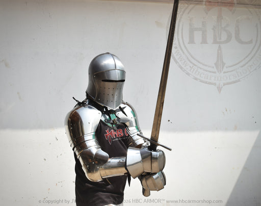A person wearing full medieval-style steel armor and a helmet holds a long sword in front of a plain light wall. A black shirt with red and white lettering is visible beneath the armor.