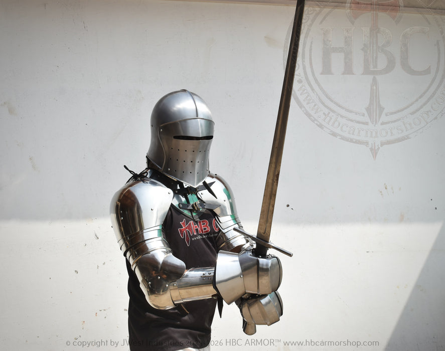 A person wearing full medieval-style steel armor and a helmet holds a long sword in front of a plain light wall. A black shirt with red and white lettering is visible beneath the armor.