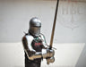 A person wearing full medieval-style steel armor and a helmet holds a long sword in front of a plain light wall. A black shirt with red and white lettering is visible beneath the armor.