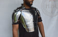 A man wearing a polished steel breastplate armor piece over a black t-shirt, shown from the chest up against a white backdrop with a faint logo.