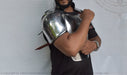 A man wearing a shiny metallic steel replica of a medieval plate armor shoulder guard, shown against a white backdrop with a faint logo watermark.