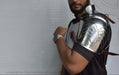 A man wearing a polished steel gladiator-style shoulder armor piece with a neck guard and leather straps, standing against a white backdrop.