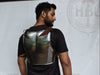 A man shown from the back wearing a metallic steel breastplate armor vest over a black t-shirt, standing in front of a white backdrop.