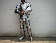 A person wearing full metallic medieval plate armor stands against a plain light wall, with the head mostly cropped out. The armor is reflective silver with dark straps and small black accents.
