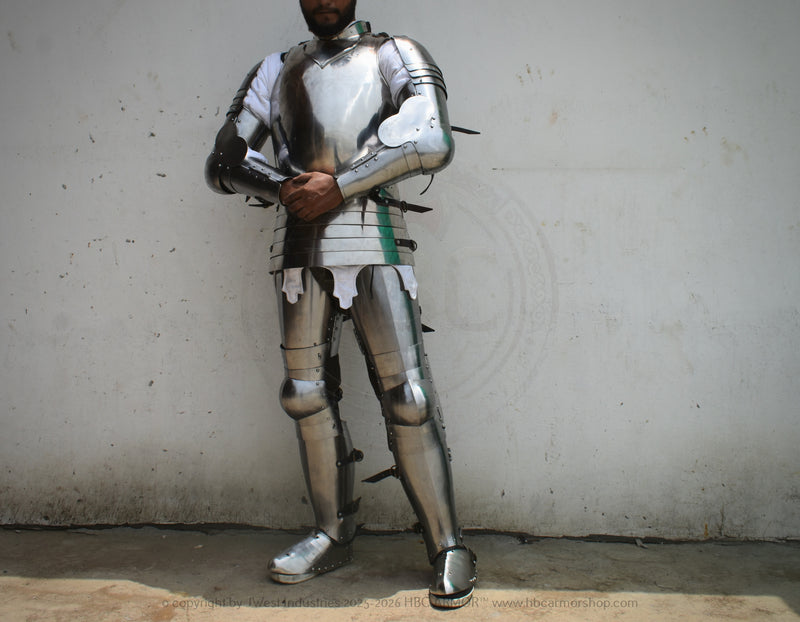 A person wearing full metallic medieval plate armor stands against a plain light wall, with the head mostly cropped out. The armor is reflective silver with dark straps and small black accents.