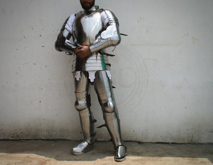 A person wearing full metallic medieval plate armor stands against a plain light wall, with the head mostly cropped out. The armor is reflective silver with dark straps and small black accents.
