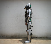 Full suit of medieval plate armor standing upright against a plain off-white wall, shown in side profile. The armor is metallic silver with black straps and small green painted accents.