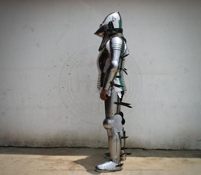 Full suit of medieval plate armor standing upright against a plain off-white wall, shown in side profile. The armor is metallic silver with black straps and small green painted accents.