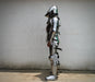 Full suit of medieval plate armor standing upright against a plain off-white wall, shown in side profile. The armor is metallic silver with black straps and small green painted accents.