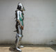 A full suit of medieval plate armor shown in profile against a plain off-white wall. The armor is primarily polished silver steel with green painted sections on the torso and upper leg, black leather straps, and a chainmail neck guard.
