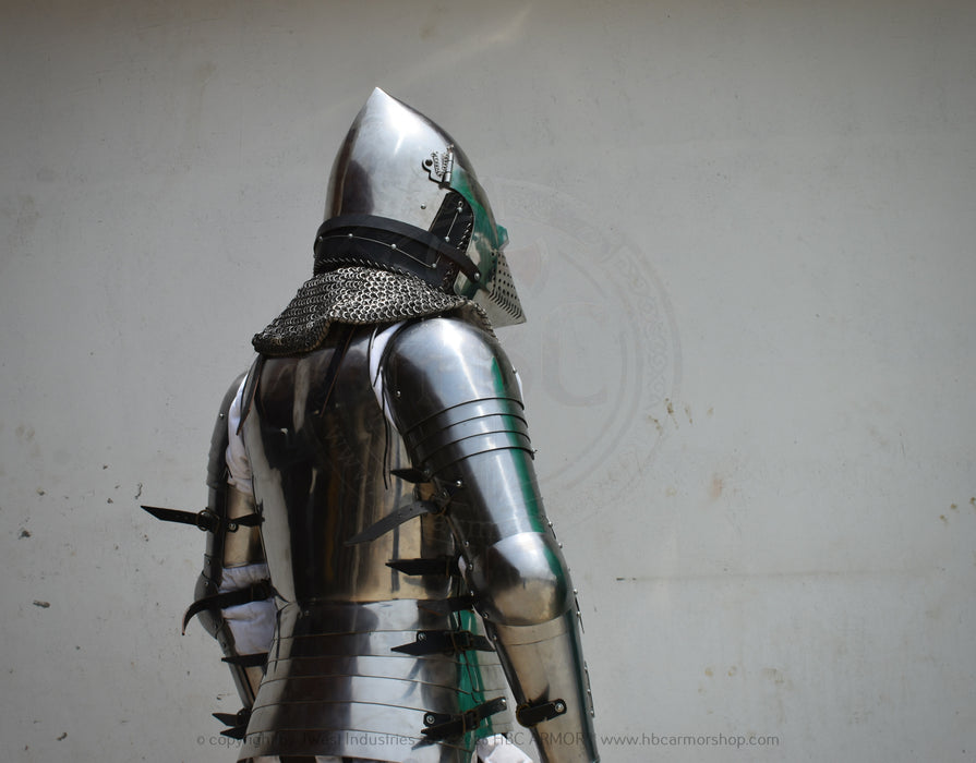 Full back view of a medieval steel plate armor suit with a pointed helmet, chainmail neck guard, and black spiked guards, shown against a plain off-white wall.