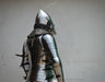 Full back view of a medieval steel plate armor suit with a pointed helmet, chainmail neck guard, and black spiked guards, shown against a plain off-white wall.