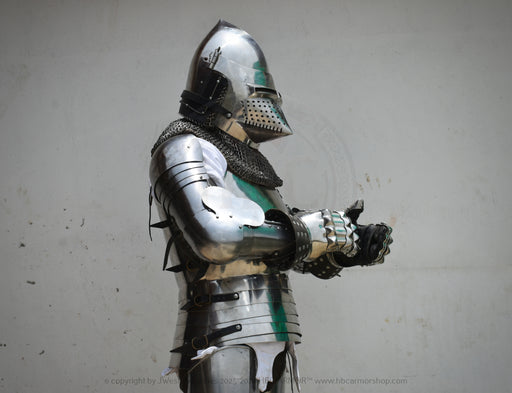 Full suit of medieval plate armor with a polished steel helmet, chainmail neck guard, articulated arm guards, and black finger guards, shown against a plain light gray wall.