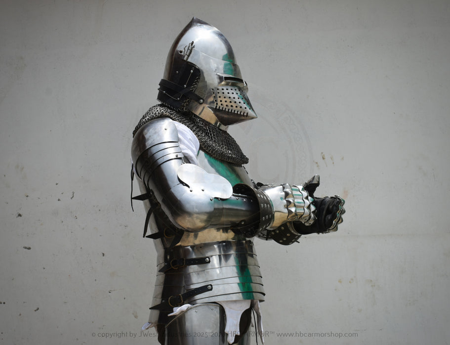 Full suit of medieval plate armor with a polished steel helmet, chainmail neck guard, articulated arm guards, and black finger guards, shown against a plain light gray wall.
