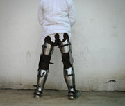 A person viewed from behind wearing a white quilted jacket and heavy metal leg armor with black straps, standing against a plain light wall.