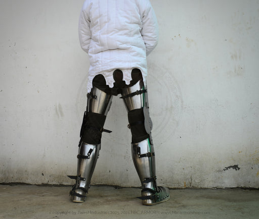 A person viewed from behind wearing a white quilted jacket and heavy metal leg armor with black straps, standing against a plain light wall.