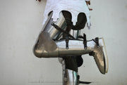 A person wearing medieval-style metal armor leg guards and a white tunic, photographed against a plain light wall.