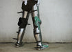 A person wearing a metallic fantasy leg armor or costume on their lower body, standing against a plain concrete wall. The armor is primarily silver with black straps and buckles, and a translucent green accent piece on the upper leg.