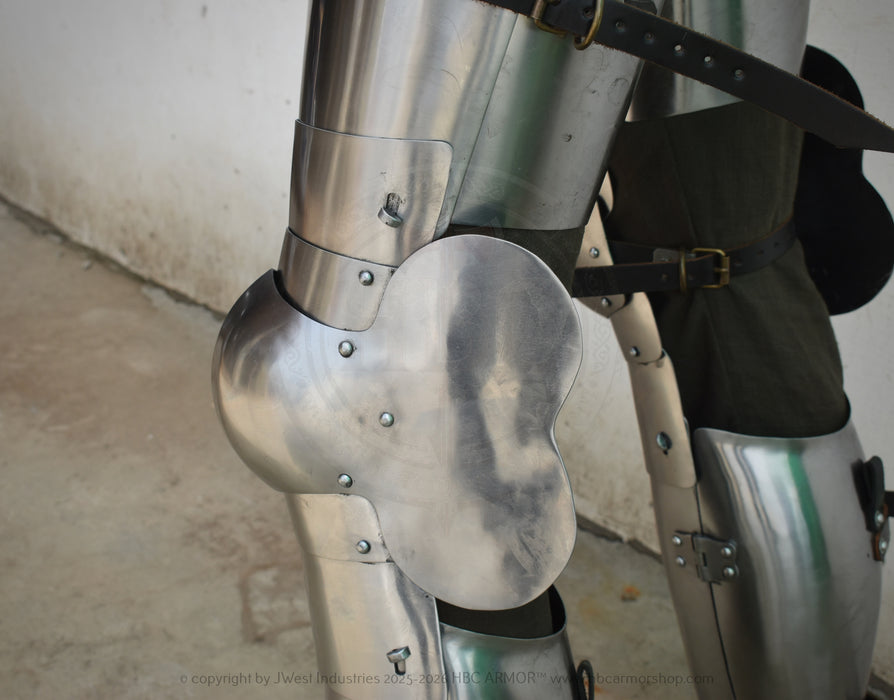 Close-up of medieval-style steel plate armor on a leg, showing polished metal plates, rivets, a curved guard, and dark straps against a concrete floor and wall.