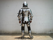 A full suit of medieval plate armor shown from the back, worn by a person standing against a plain off-white wall.