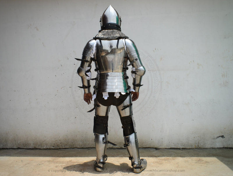 A full suit of medieval plate armor shown from the back, worn by a person standing against a plain off-white wall.