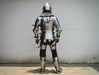 A full suit of medieval plate armor shown from the back, worn by a person standing against a plain off-white wall.