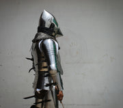 A person wearing medieval-style steel plate armor and a helmet, shown in profile against a plain light gray wall. The armor includes polished steel sections, chainmail neck protection, and dark straps.